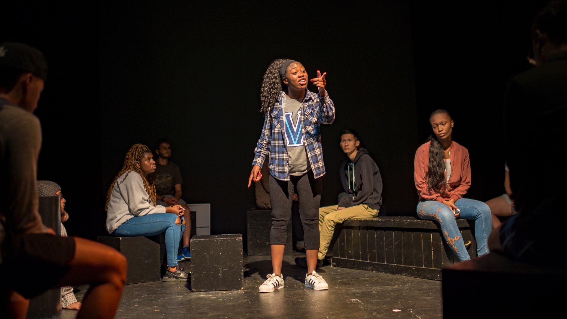 A female student standing speaking to a crowd in Vasey Theatre with several male and female students seated listening
