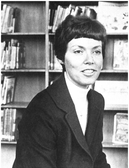 A black and white photo of a white woman with short hair in a suit, near a bookcase