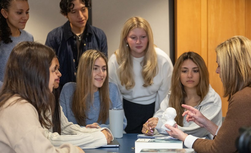 Students and professor discuss an artifact in a classroom. image of students and teacher seated around table