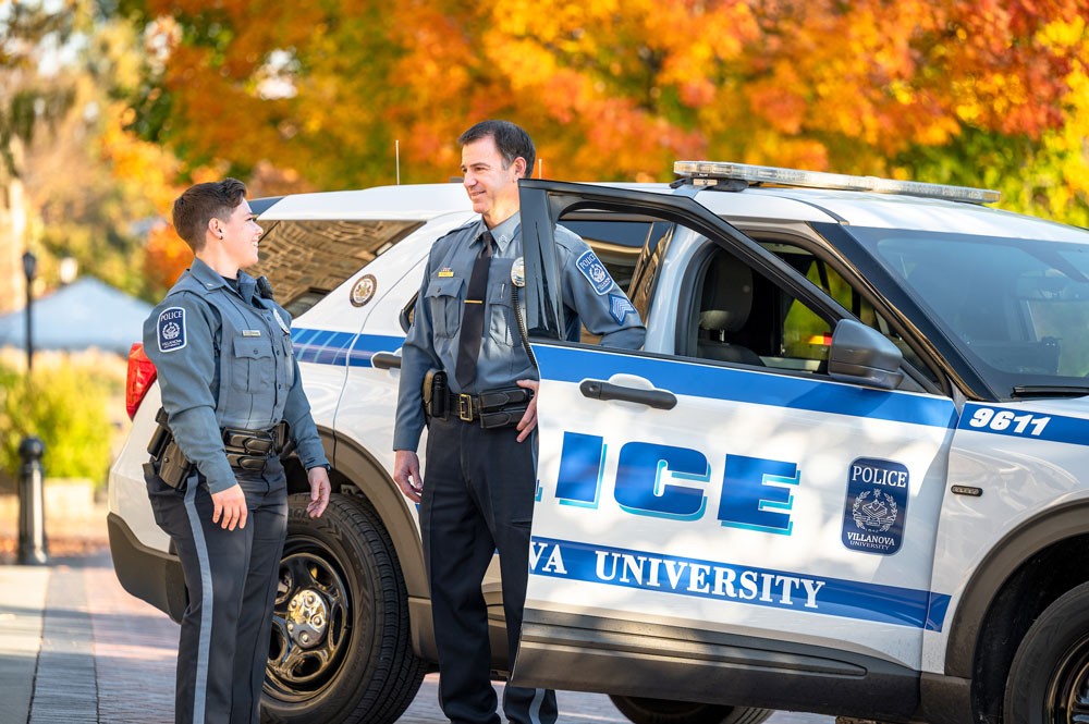A male and a female public safety officer talk outside a patrol car.