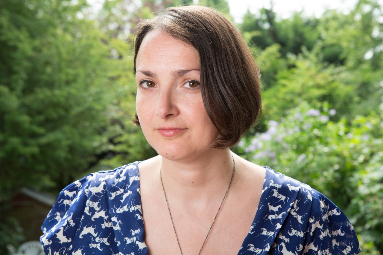 Photo of Hannah Khalil from the shoulders up. She is a white woman with chin-length brown hair. She is wearing a blue v-neck top and is looking at the camera.